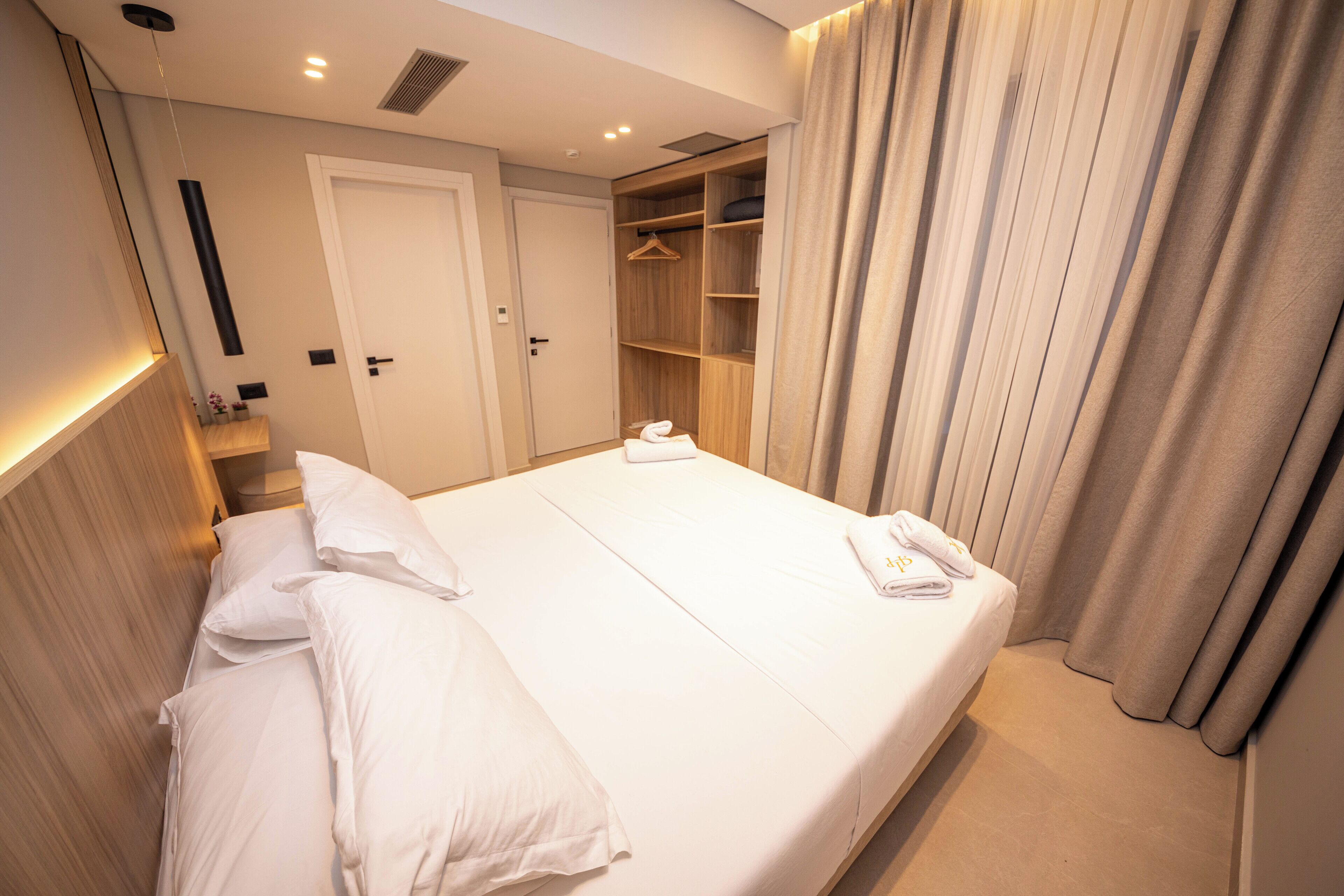 A neatly made bed with white linens, a wooden headboard, a built-in wardrobe with hanging space, and a door leading to a bathroom.
