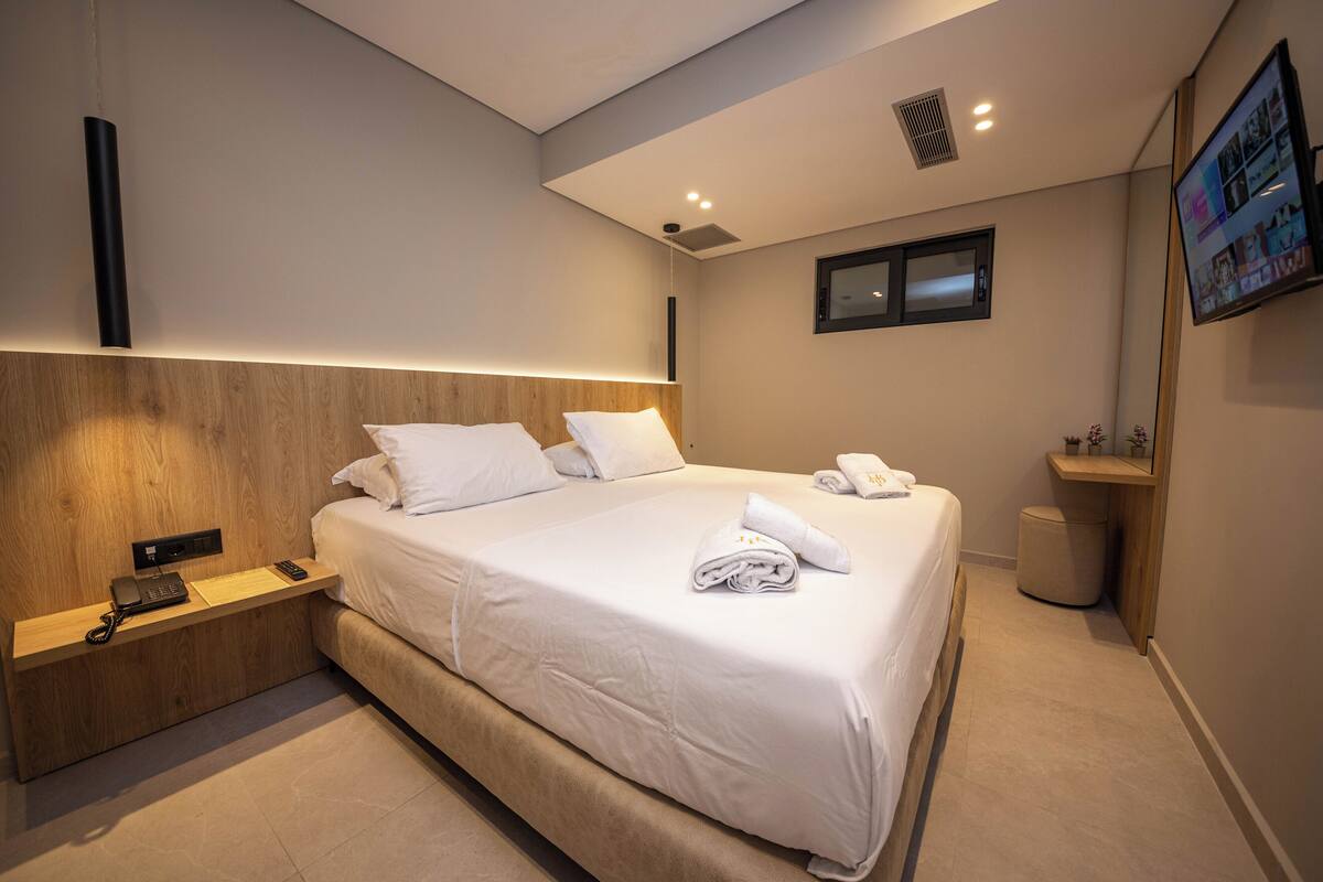 Une chambre d’hôtel moderne avec un grand lit, une table de chevet avec un téléphone, un téléviseur à écran plat fixé au mur et une petite étagère en bois.