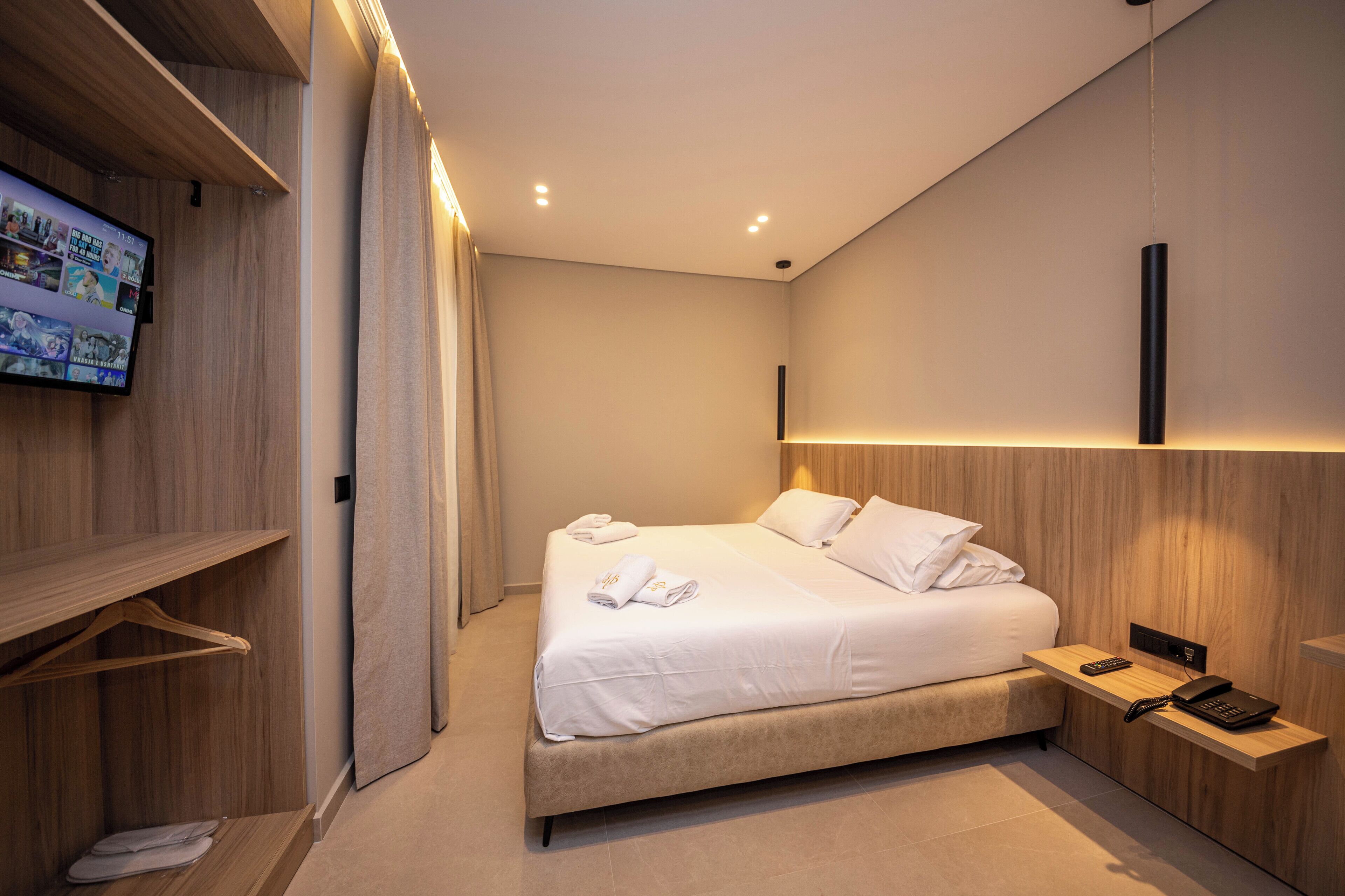Habitación de hotel moderna con una cama grande, una mesita de noche con teléfono, un televisor montado en un panel de madera y un armario de madera.