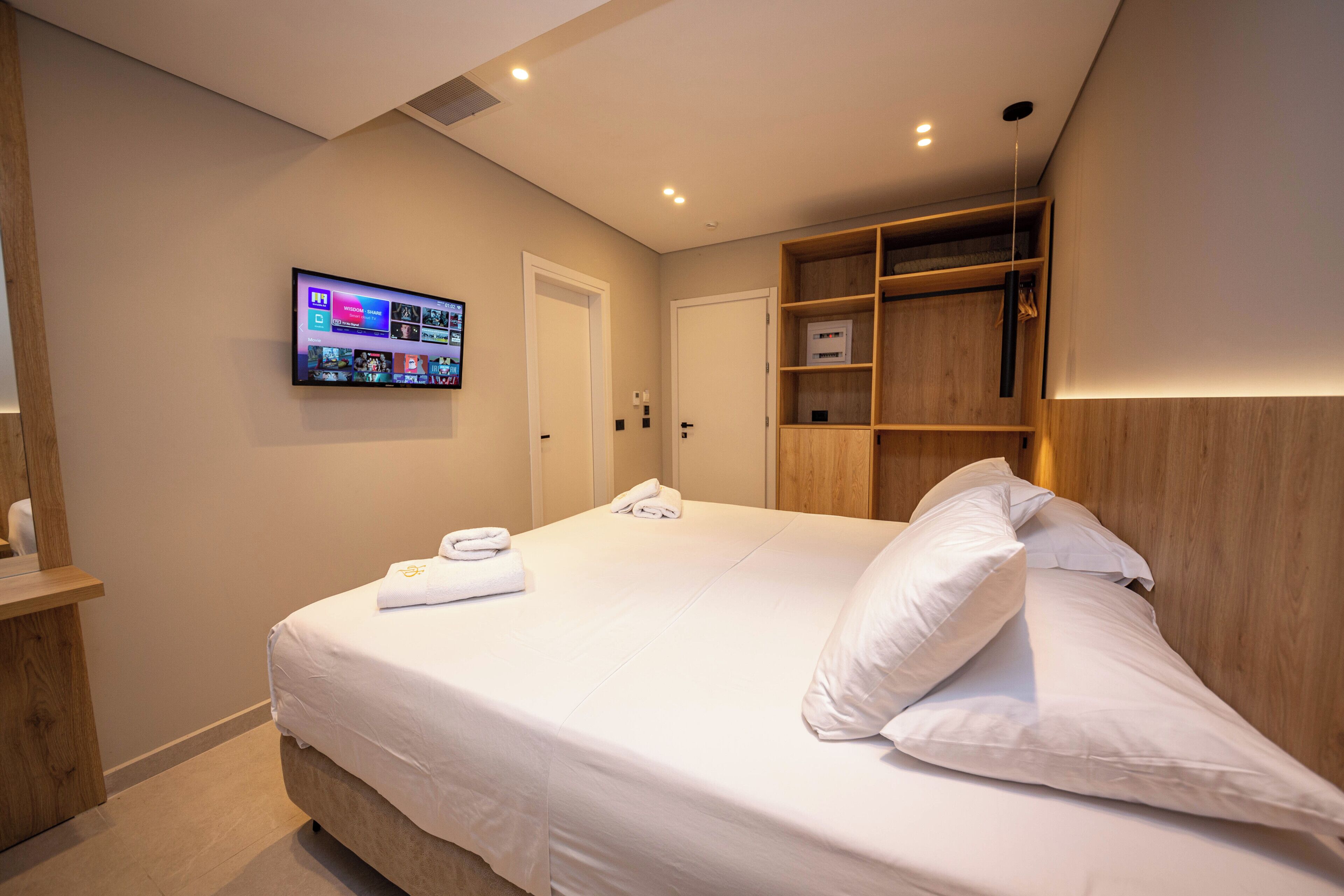 Una cama bien hecha con sábanas y almohadas blancas, un televisor de pantalla plana en la pared y un estante de madera con algunos objetos.