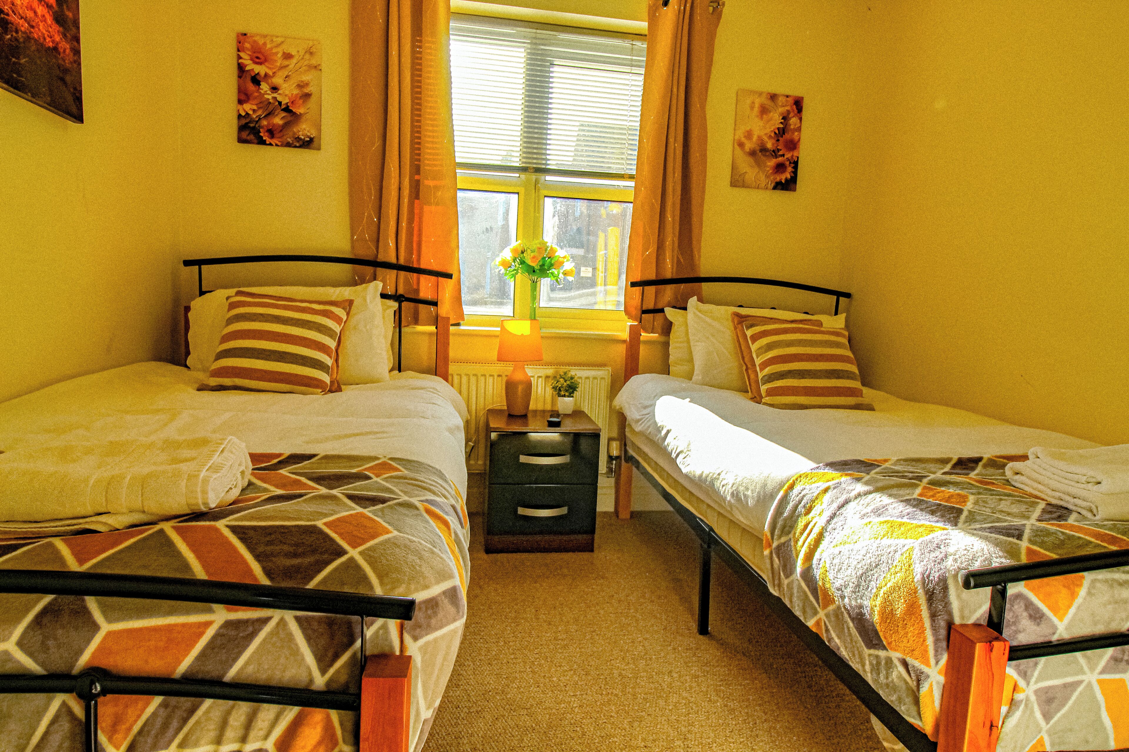 Habitación doble con dos camas, una ventana con cortinas, una mesita de noche con lámpara y un jarrón con flores.