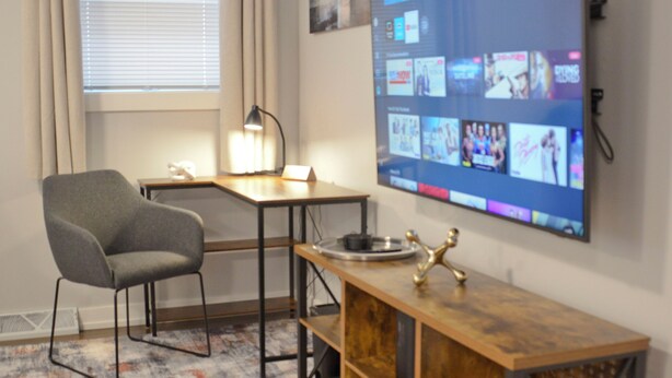 A modern living room with a flat-screen TV mounted on the wall, a wooden coffee table, a grey armchair, and a desk with a lamp.