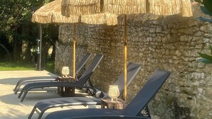 A row of sun loungers under a thatched umbrella, with a stone wall in the background. - Le Château de Leugny (Plaine-et-Vallées)
