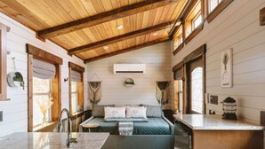 Interior acogedor de una cabaña con techo de madera, una cama con manta verde, un lavamanos y una pequeña cocina.