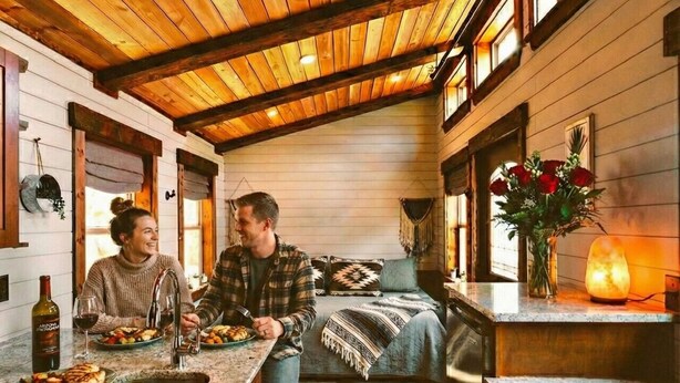 Interior acogedor de una cabaña con techo de madera, una zona de cocina con encimera de granito y una cama con manta estampada.