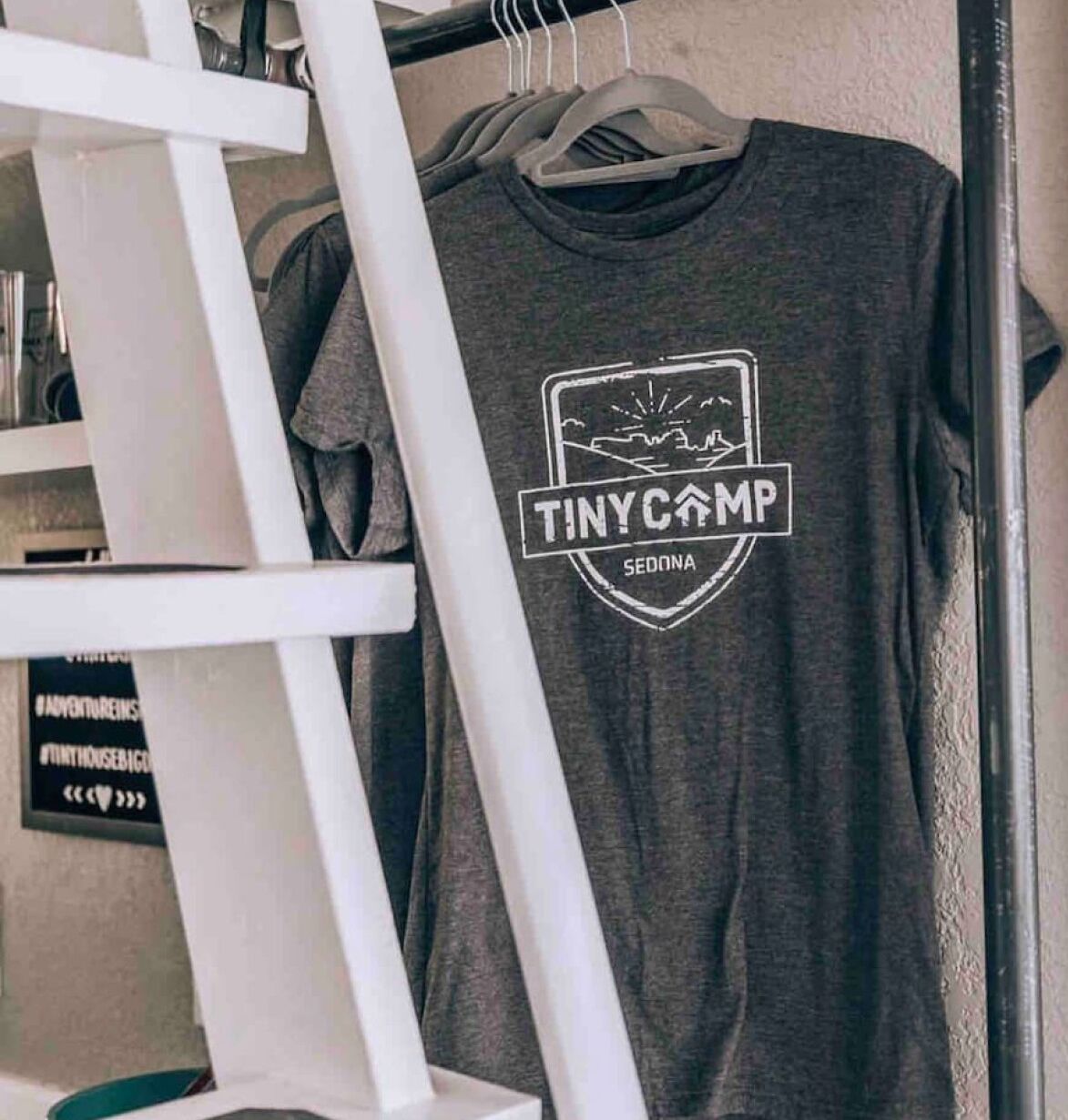 En grå t-shirt med texten ”TINY CAMP” och ”SEDONA” på, hängande på en klädställning.