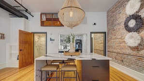 A modern kitchen with a central island, wooden stools, and a hanging light fixture.