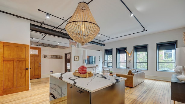 A spacious room with a large island, wooden flooring, a hanging light fixture, and large windows.