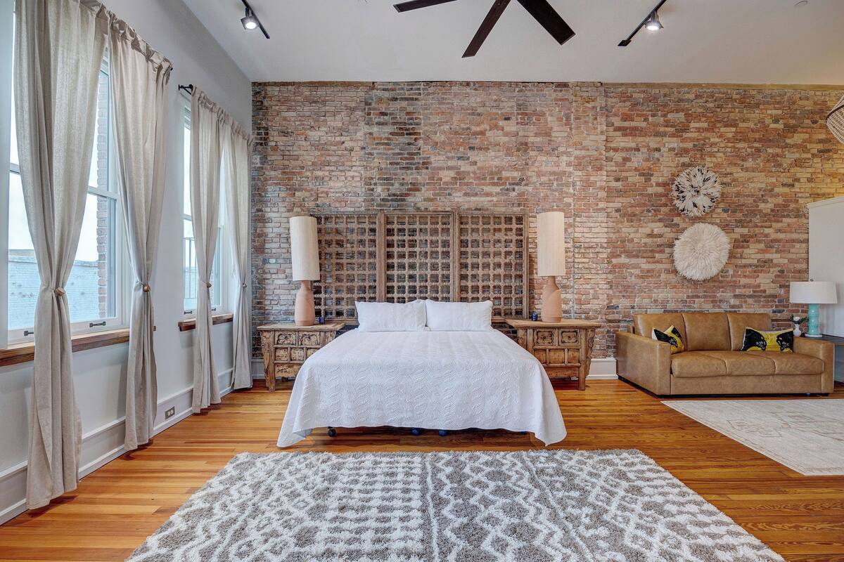Un dormitorio con pared de ladrillo, una cama grande, un sofá y una alfombra estampada.