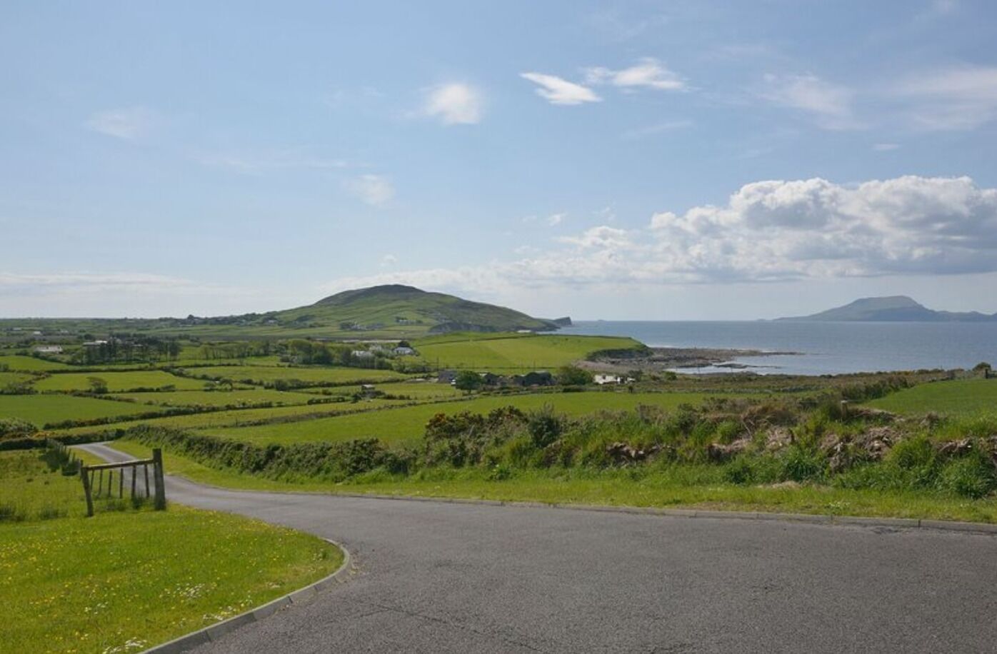 Un paysage rural avec une route qui serpente à travers des champs verdoyants, un petit banc et une vue lointaine sur la mer et les collines.