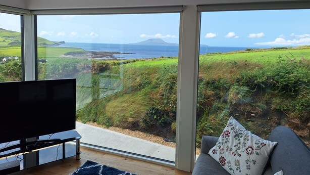 Una sala moderna con un televisor grande, un sofá con un cojín floral y vistas a un paisaje costero a través de ventanales.
