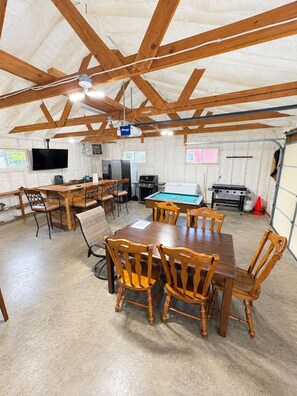 A spacious indoor area with a pool table, wooden dining table, and chairs, featuring a high ceiling with exposed beams and a television mounted on the wall.