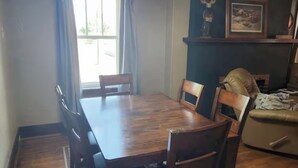Un coin repas avec une table et des chaises en bois, un tapis à motifs, une fenêtre avec des rideaux et une étagère murale ornée d’un tableau encadré.