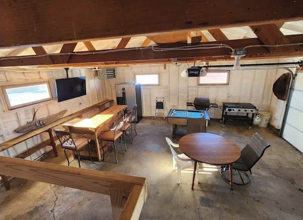 A rustic, open-plan space with a wooden dining table, bar area, pool table, and a grill.