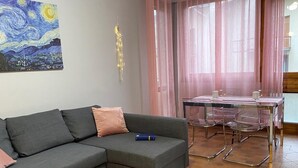 A living room with a grey sofa, a painting on the wall, a dining area with a table and chairs, and a window with sheer curtains.
