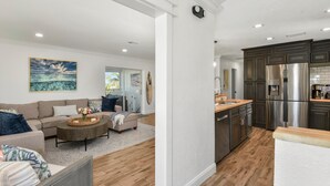 A modern living room with a sofa, coffee table, and artwork. A kitchen with stainless steel appliances and wooden countertops is visible. - Coral Cove (Fort Myers Beach)