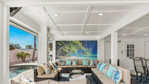 A spacious living area with a large sofa, a coffee table, and a view of a swimming pool and palm trees. - Coral Cove (Fort Myers Beach)