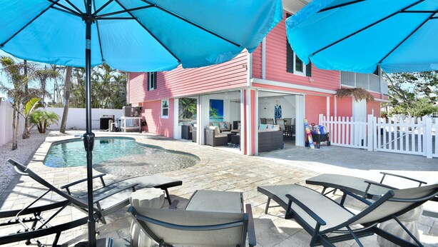 Een zwembadterras met ligbedden en parasols, een zwembad en een roze gebouw met terras.