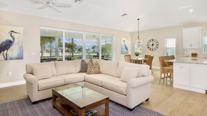 A spacious living room with a large L-shaped beige sofa, a glass coffee table, and a dining area with wicker chairs. - Holiday Everyday (Fort Myers Beach)