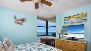 A bedroom with a bed, a desk, a TV, a ceiling fan, a window with a view, and a wall with a painting. - Lovers Key Beach Club #303 (Fort Myers Beach)