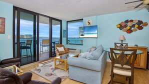 A living room with a sofa, coffee table, and chairs, a dining table with chairs, a lamp, and a wall with a painting and a decorative wall hanging. - Lovers Key Beach Club #303 (Fort Myers Beach)