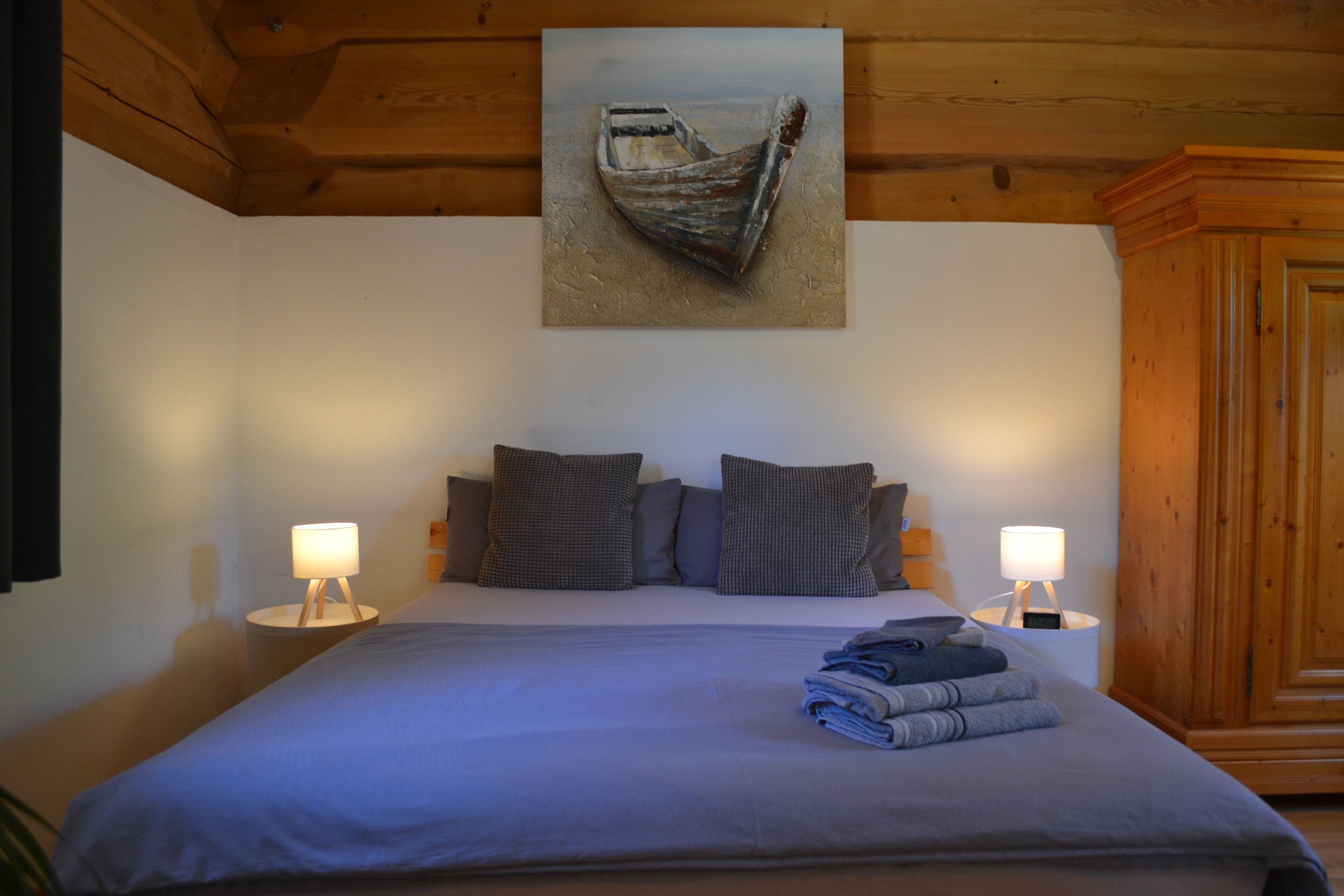 Una cama bien hecha con una colcha azul, dos lámparas de noche y un cuadro de un barco en la pared.
