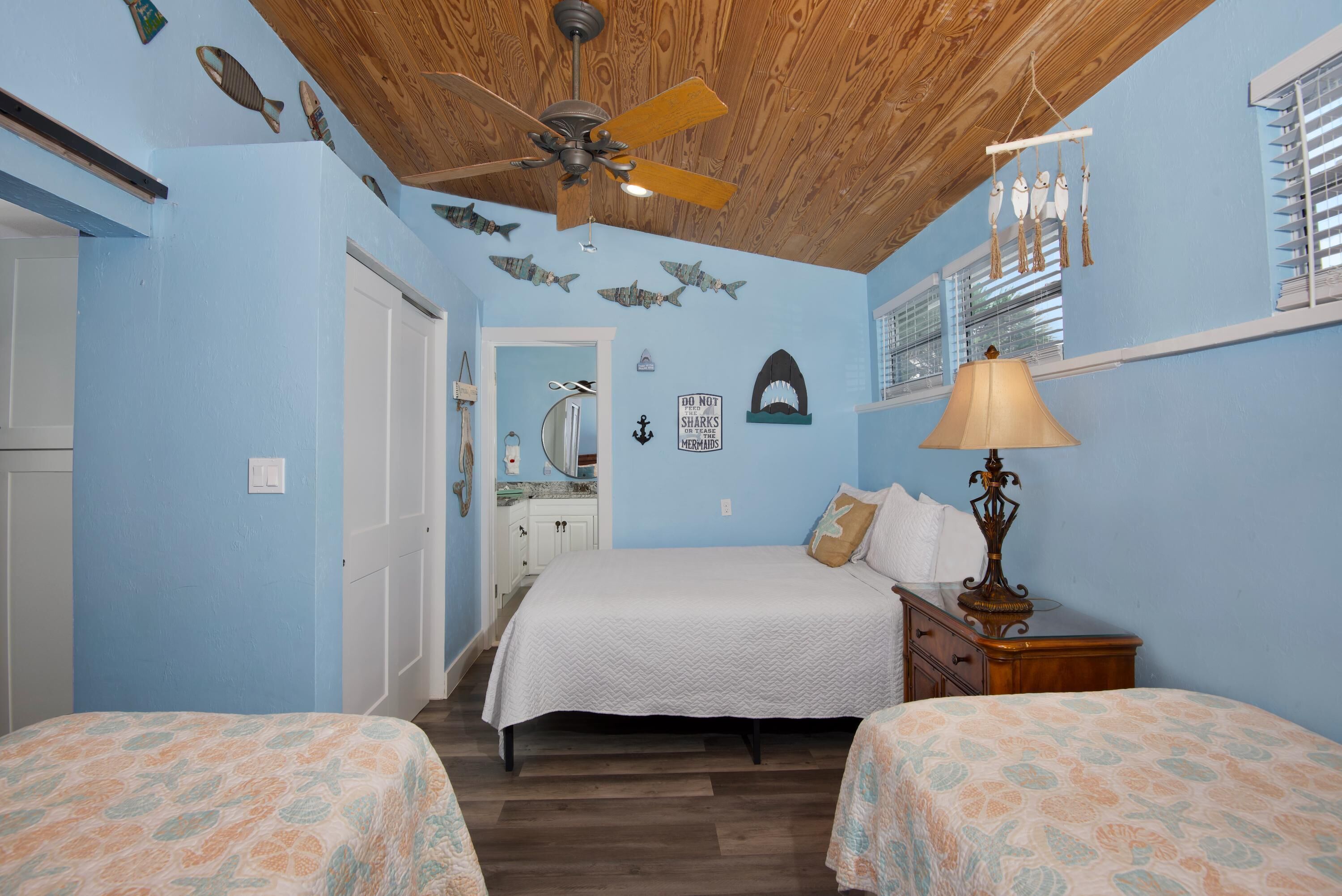 Un dormitorio con dos camas, un ventilador de techo, una mesita de noche con lámpara y decoración en la pared.