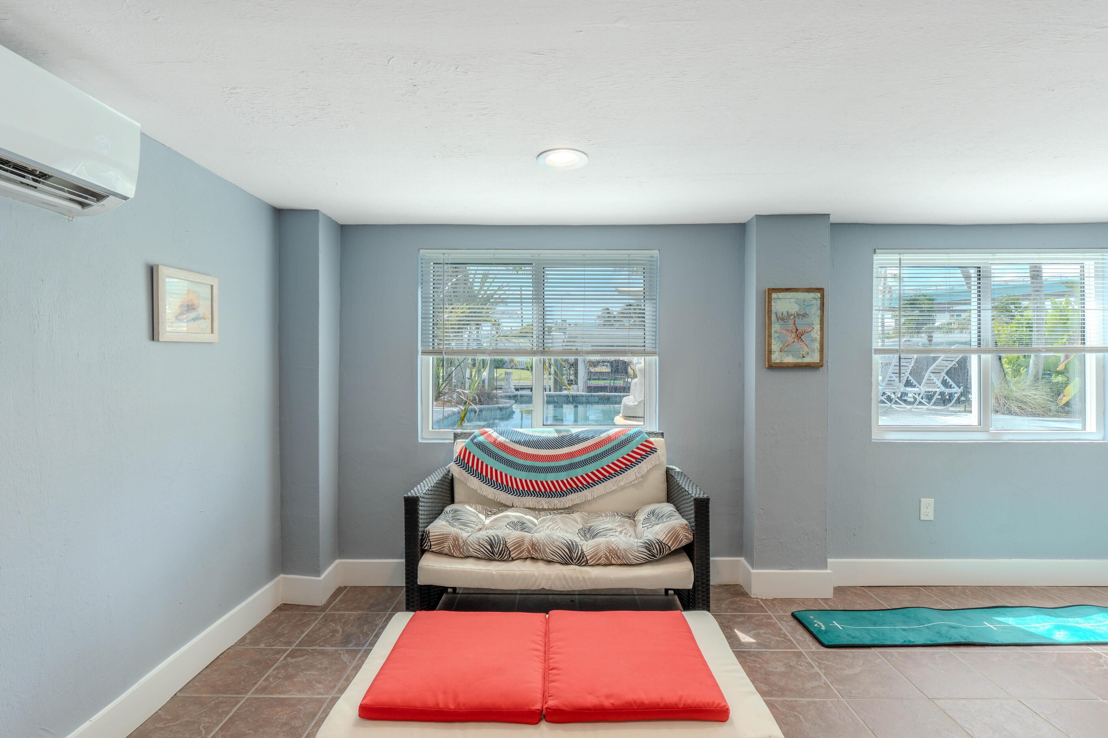 A room with a cushioned bench, a chair with a patterned throw, and two windows with blinds.
