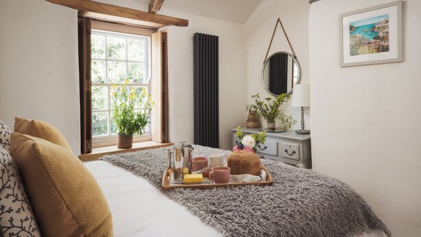A bedroom with a large window, a bed with a fluffy grey blanket, a bedside table with a lamp, and a tray with breakfast items. - Cornwall Getaway for two - The Loft (Saint Kew)