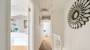Ein Korridor mit Holzboden, weißen Wänden und einem weißen Geländer. An der Wand befindet sich ein dekorativer Spiegel und eine Tür zu einem Schlafzimmer.