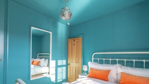 Ein Schlafzimmer mit türkisfarbenen Wänden, einem weißen Bett mit orangefarbenen Kissen, einer Holztür und einer Pendelleuchte.