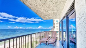 Een balkon met uitzicht op de oceaan, twee gestreepte lounge stoelen en een glazen deur die naar binnen leidt.