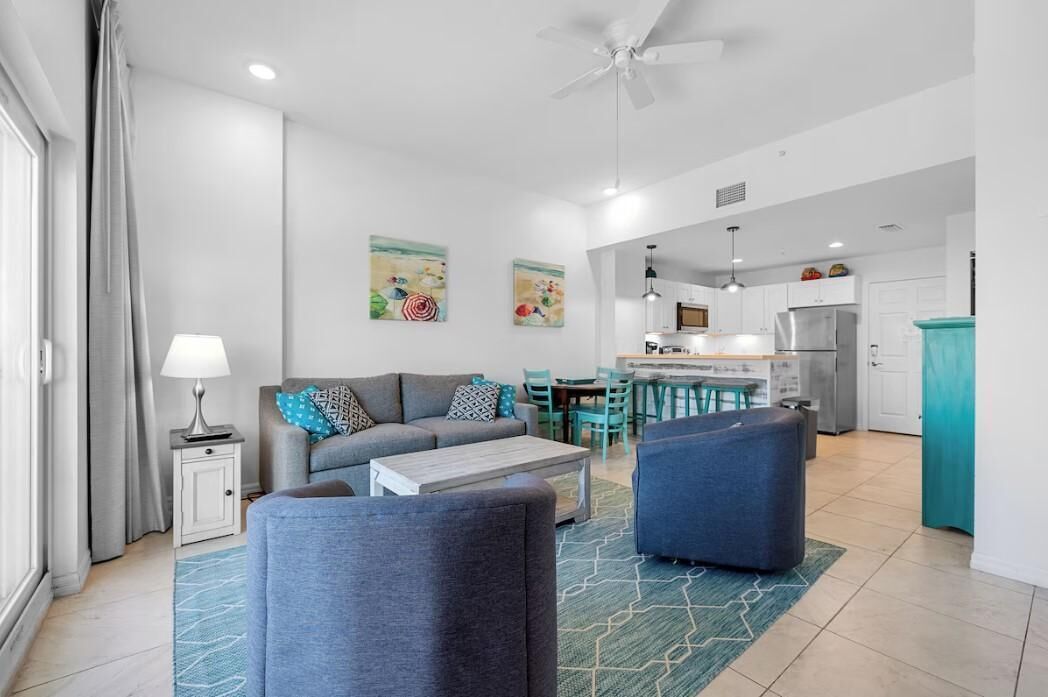 A modern living room with a sofa, coffee table, and ottoman, a dining area with chairs, and a kitchen with a refrigerator and cabinets.
