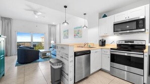 A modern kitchen with stainless steel appliances, a dishwasher, and a dining area with a blue sofa and a view of the outdoors. - Madeira Bay Resort I 1402 (Madeira Beach)