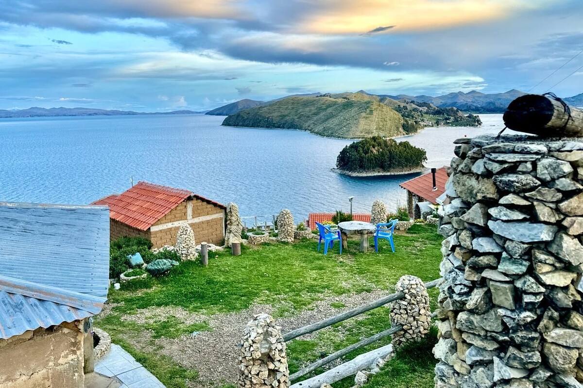 A lakeside property with a stone wall, a small building with a red roof, and a grassy area with blue chairs.