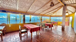 A restaurant with wooden tables and chairs, large windows with curtains, and a view of the sea. - Kalluchi Ecolodge (La Paz)