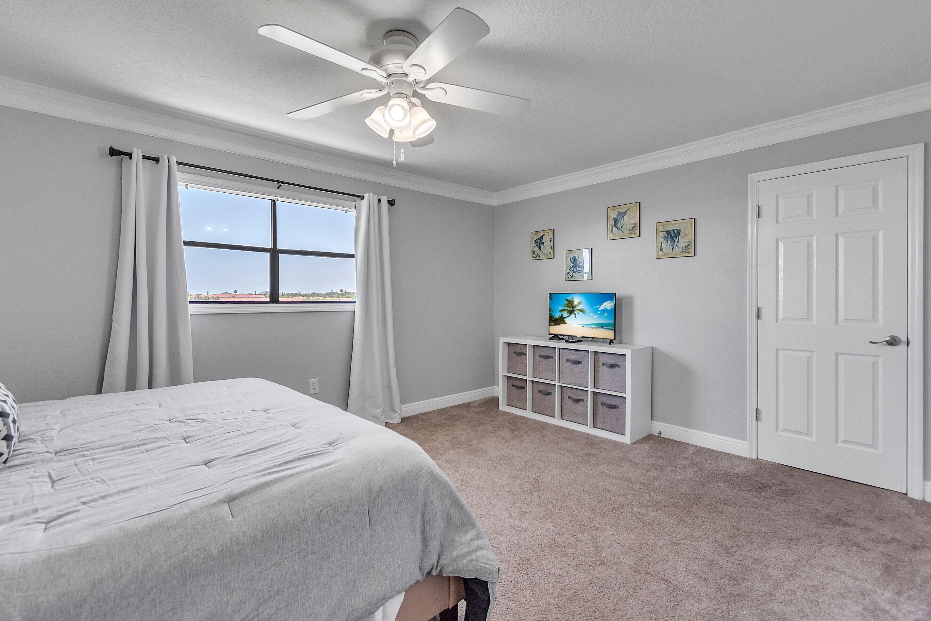 Un dormitorio con cama, ventilador de techo, televisor sobre un mueble bajo y una ventana con cortinas.
