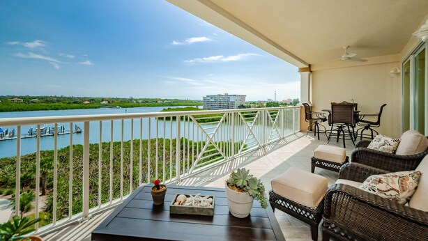 A balcony with wicker furniture, a table with decorative items, and a view of a river and distant buildings. - Dolphin Key 504 - Premier (Indian Shores)