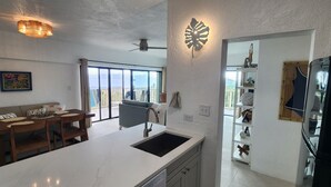 A modern kitchen with a large island, a dining area with a sofa and chairs, and a view through large windows. - NEWLY RENOVATED! 1BR/1BA Beautiful Condo @Point Pleasant/Great views/3 Pools! (St Thomas)