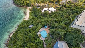 An aerial view of a coastal property with a swimming pool surrounded by dense greenery, a beach, and a road. - NEWLY RENOVATED! 1BR/1BA Beautiful Condo @Point Pleasant/Great views/3 Pools! (St Thomas)