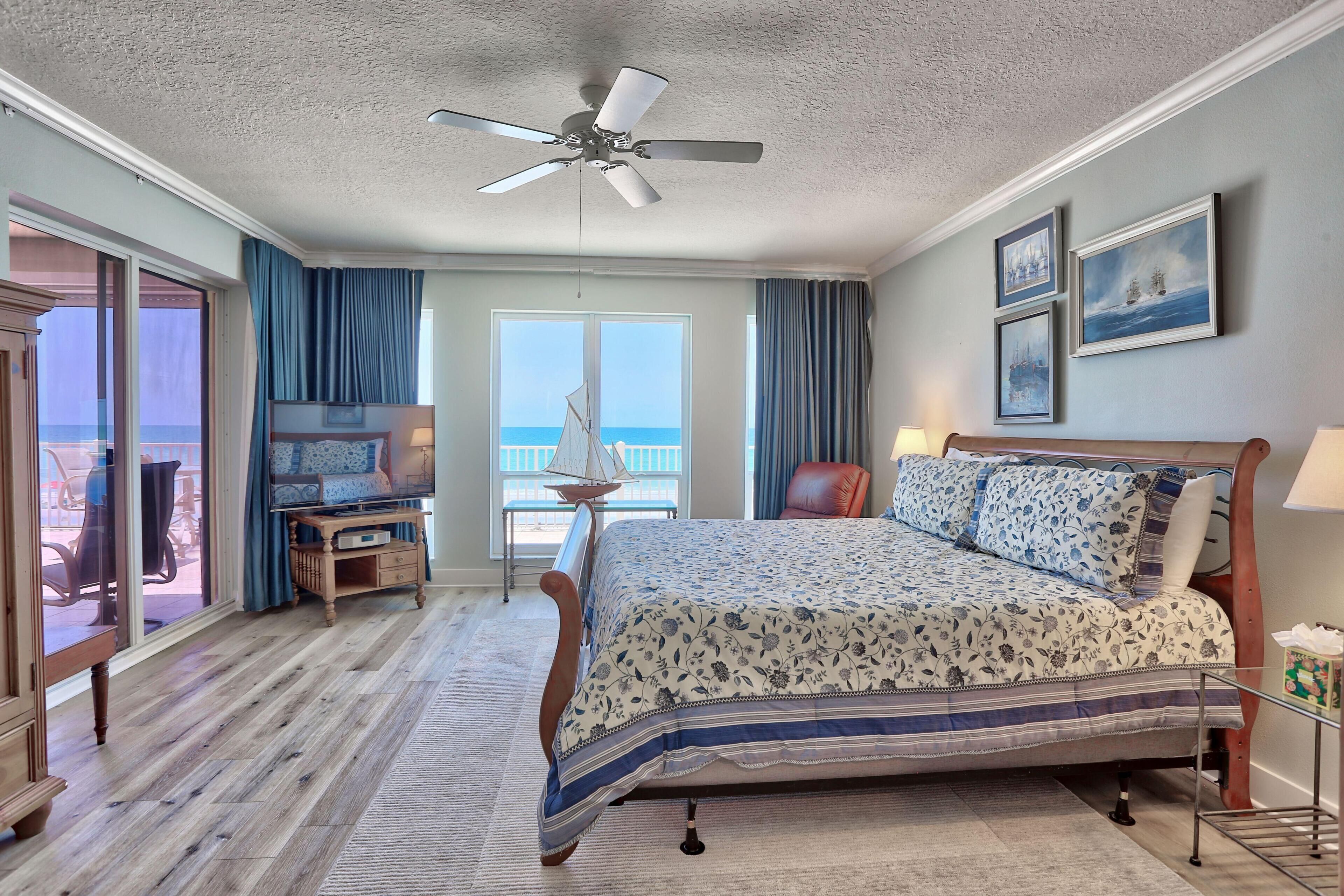 A bedroom with a bed, a ceiling fan, a window with a view of the ocean, a chair, and a small table.