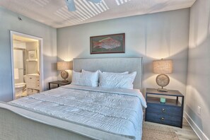 Une chambre avec un grand lit, deux tables de chevet avec des lampes, une œuvre d’art représentant un poisson encadrée au mur, et une salle de bain visible  à travers une porte ouverte.