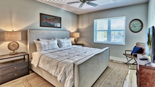 A bedroom with a bed, two bedside tables, a ceiling fan, a window with blinds, and a framed picture on the wall. - Tides 1-211 (North Redington Beach)