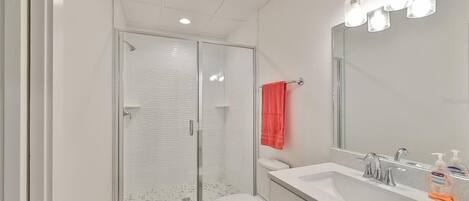 Un baño moderno con cabina de ducha de vidrio, inodoro blanco, lavabo con espejo y una toalla roja colgada en la pared.