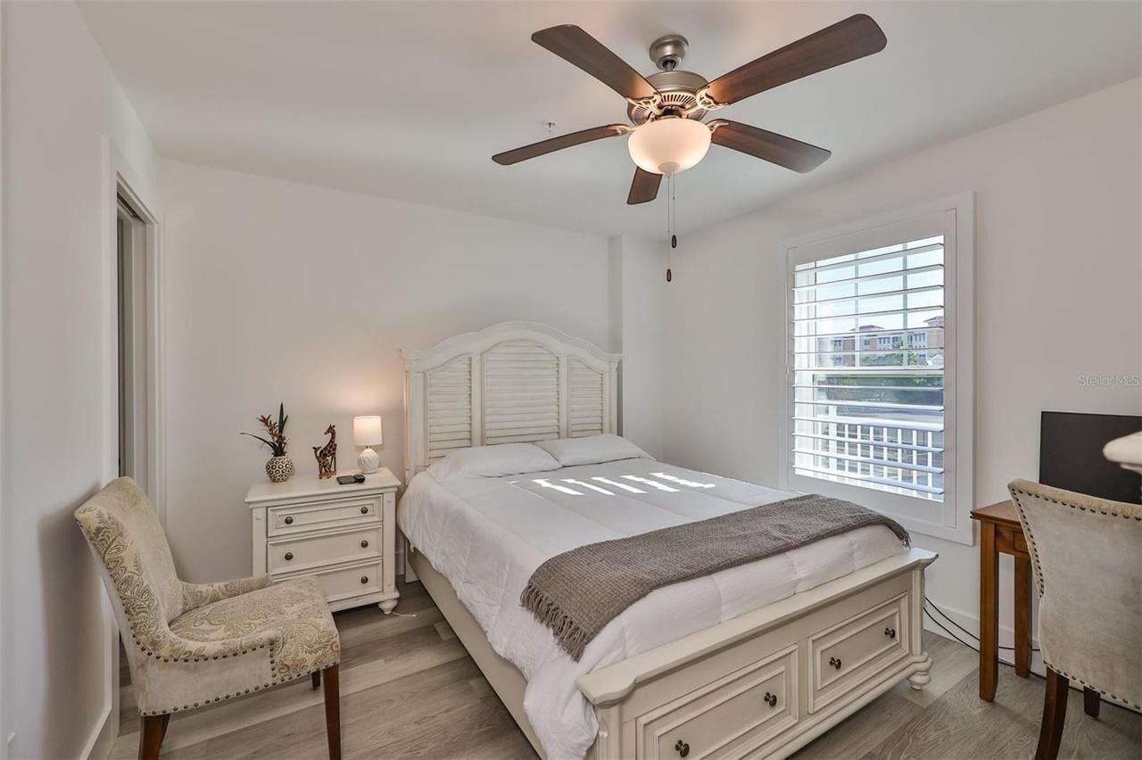 Une chambre à coucher comprenant un lit, une chaise, une table de chevet, un ventilateur de plafond et une fenêtre avec des stores.