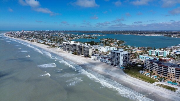 An aerial view of a coastal city with a beachfront hotel featuring multiple floors, a swimming pool, and a marina in the background. - Madeira Del Mar 301 (Madeira Beach)