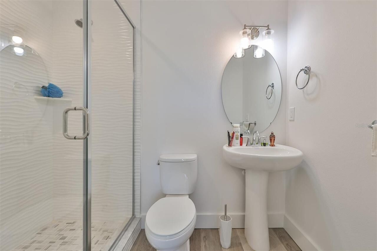 Un baño moderno con cabina de ducha de vidrio, inodoro blanco, lavamanos pedestal y espejo redondo.