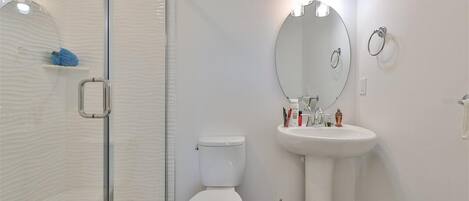 Un baño moderno con cabina de ducha de vidrio, inodoro blanco, lavamanos pedestal y espejo redondo.