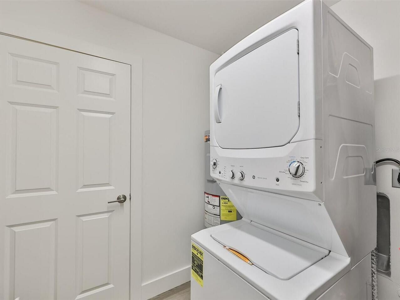 A laundry room with a stacked washer and dryer, a white door, and a caution sign on the dryer.