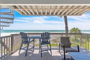 A deck with two Adirondack chairs, a small table, and a wicker chair, overlooking a beach with a clear sky. - Pelican Pointe 4C (Indian Shores)